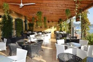 Café al aire libre con mesas y sillas, techo de madera y plantas colgantes.