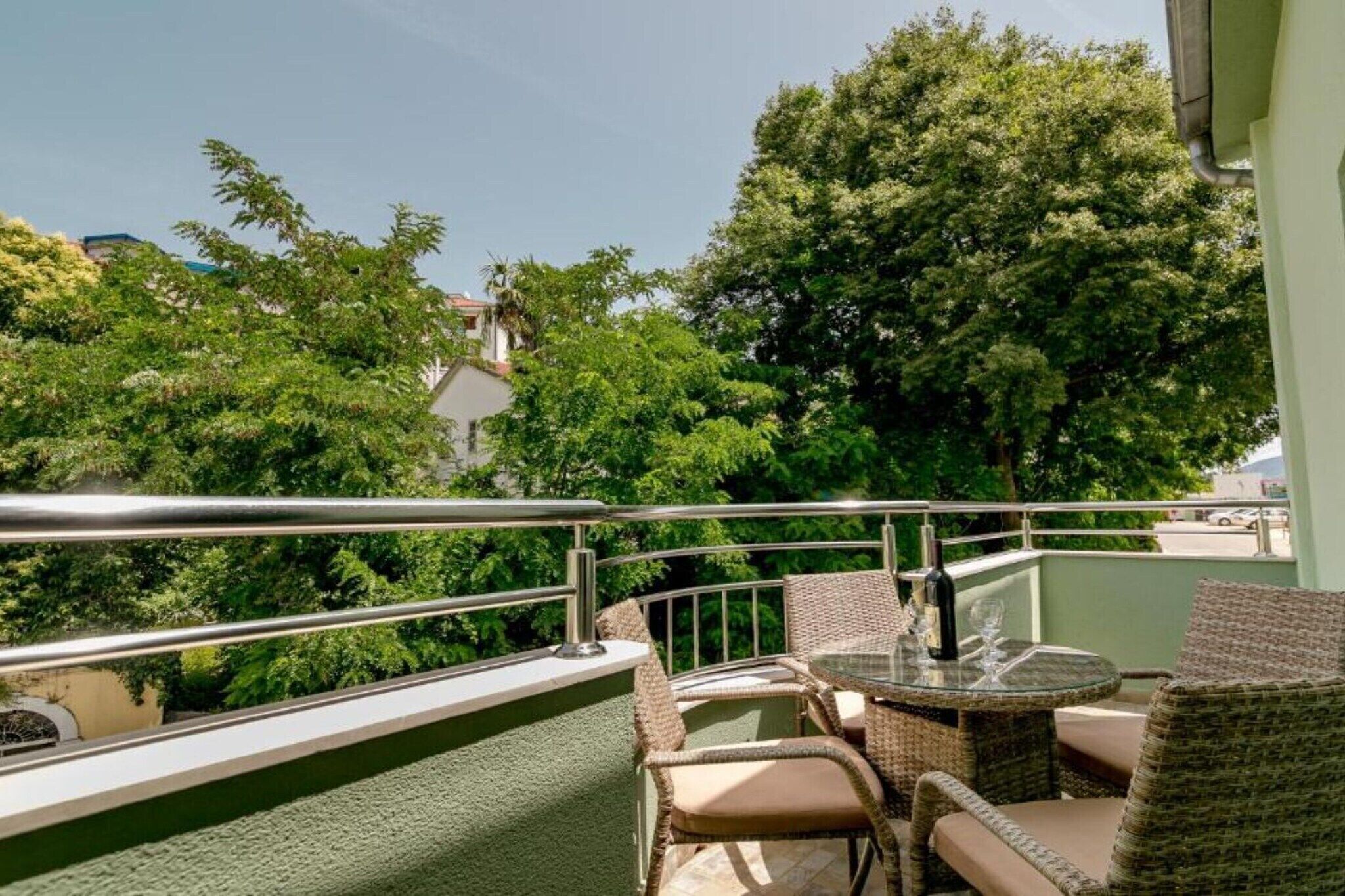 Un balcon agrémenté d’une table en verre et de chaises en osier, entouré d’une végétation luxuriante et muni d’une rambarde en métal.