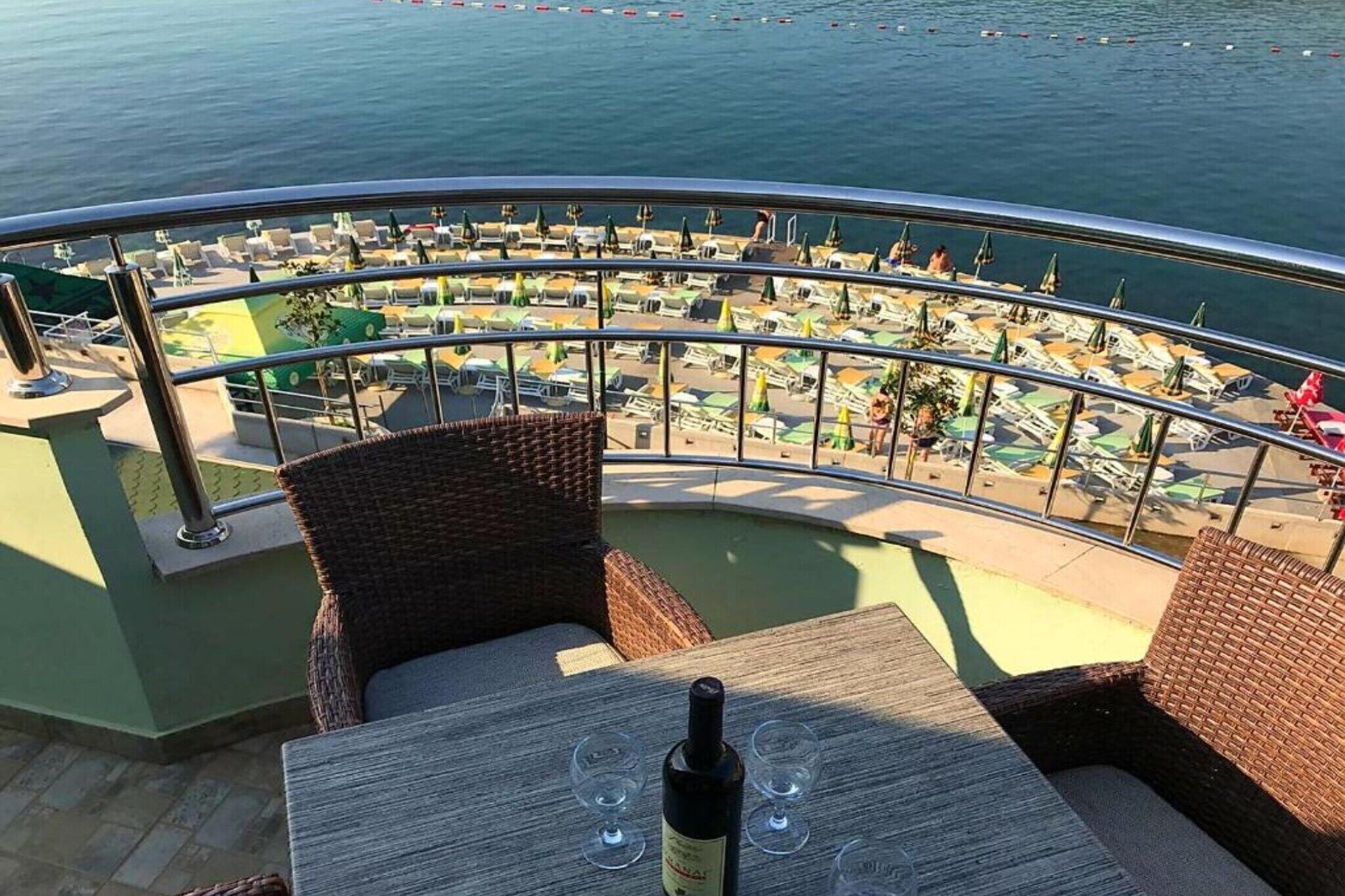 A balcony with wicker furniture, a table with a bottle and glasses, and a view of a beach with umbrellas and lounge chairs.