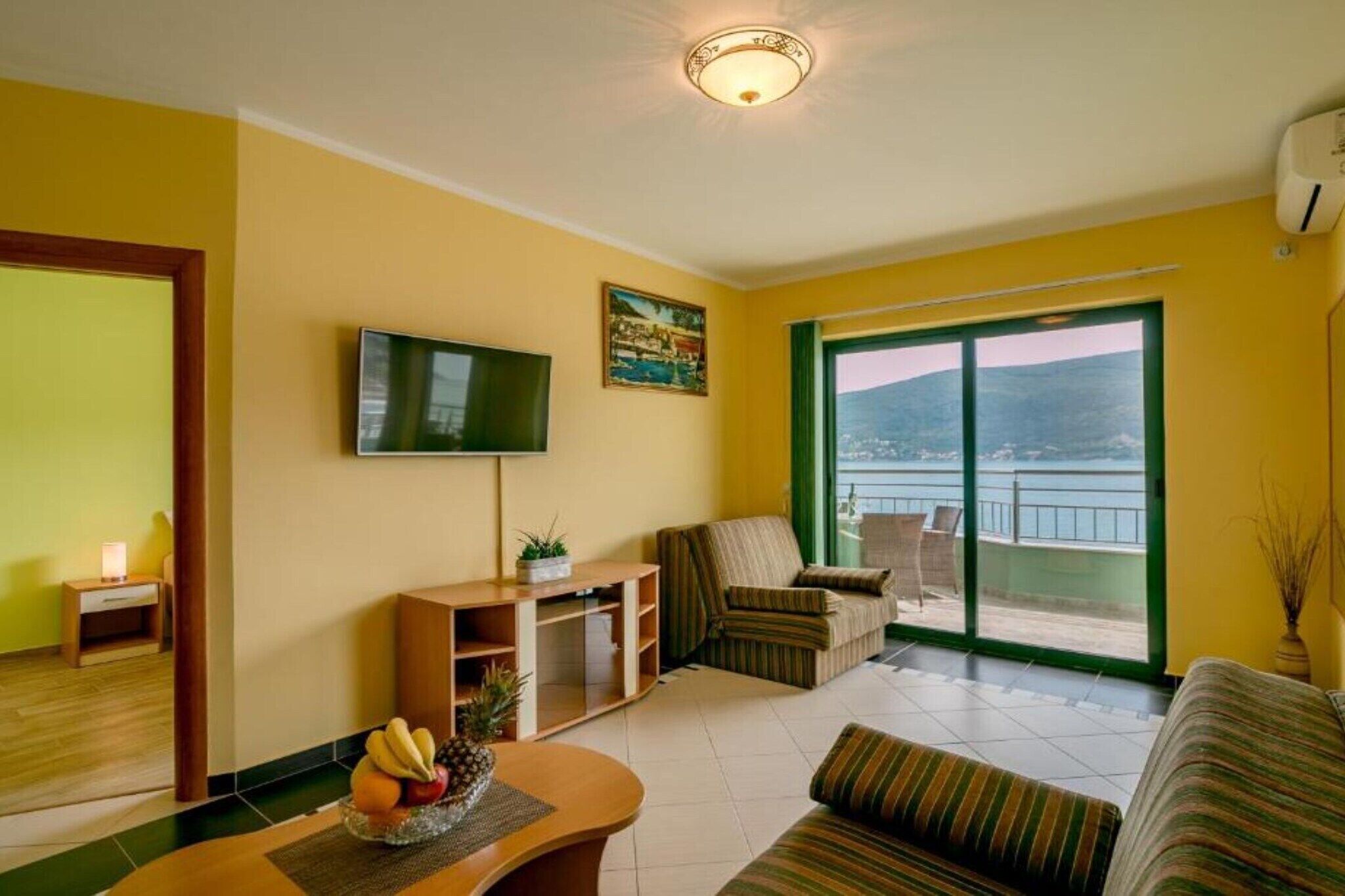 Un salón con televisión de pantalla plana, un sofá, una mesa de comedor con fruta y vistas al mar a través de un ventanal amplio.