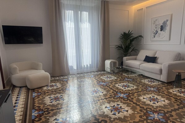 Un salón moderno con piso estampado, un sofá blanco, una mesa de centro de vidrio y un ventanal con cortinas traslúcidas.