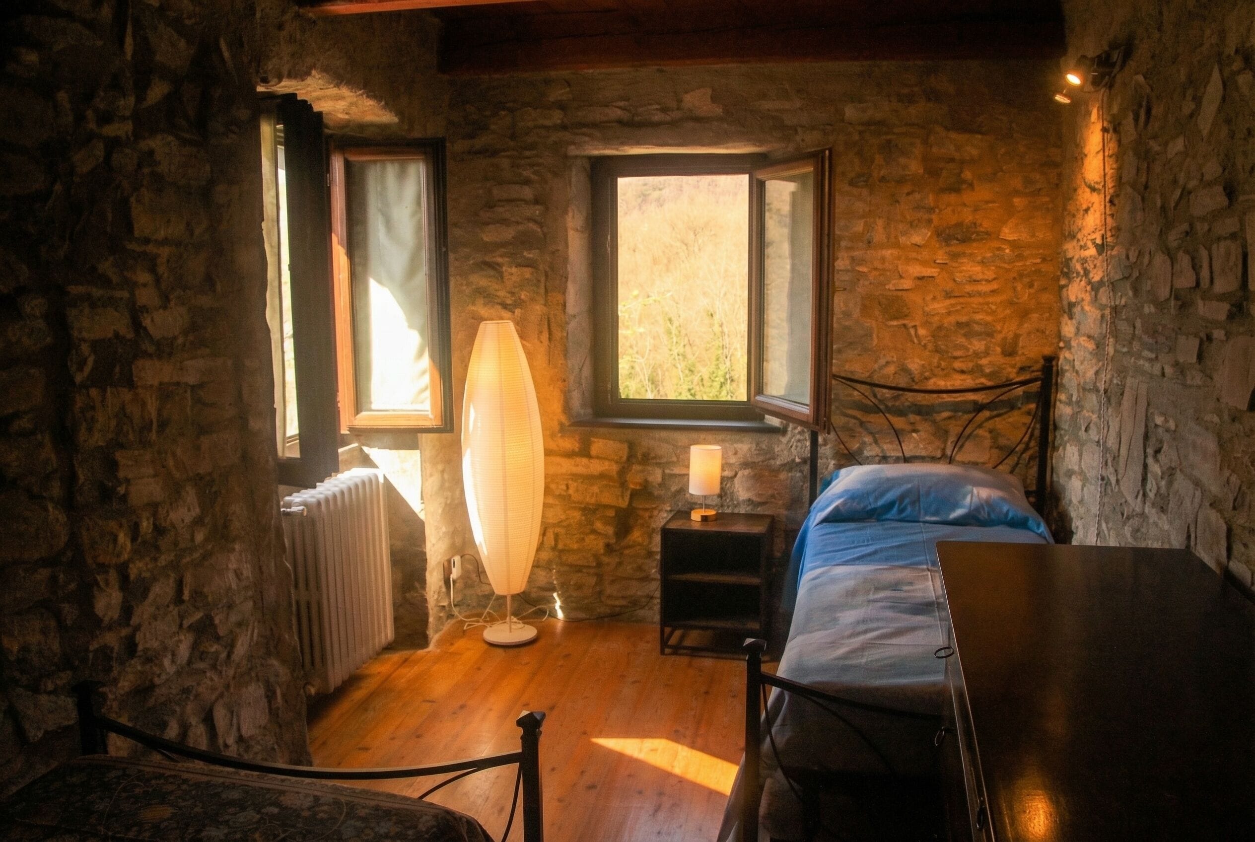 Una stanza con pareti in pietra, un letto con materasso blu, un comodino con una lampada e un termosifone.