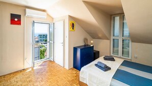 Ein Schlafzimmer mit Bett, Kommode, einem Fenster mit Jalousien, einer Tür und Blick ins Freie.