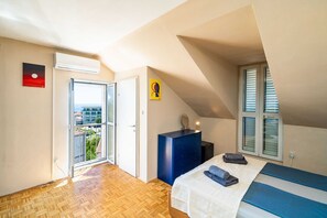 Ein Schlafzimmer mit Bett, Kommode, einem Fenster mit Jalousien, einer Tür und Blick ins Freie.