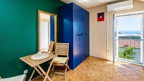 Ein Zimmer mit Holzboden, einem Tisch mit Stuhl, einem großen blauen Kleiderschrank, einem Spiegel mit Goldrahmen und einem Fenster mit Blick auf Gebäude und Grünflächen.