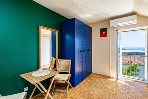 Ein Zimmer mit Holzboden, einem Tisch mit Stuhl, einem großen blauen Kleiderschrank, einem Spiegel mit Goldrahmen und einem Fenster mit Blick auf Gebäude und Grünflächen.