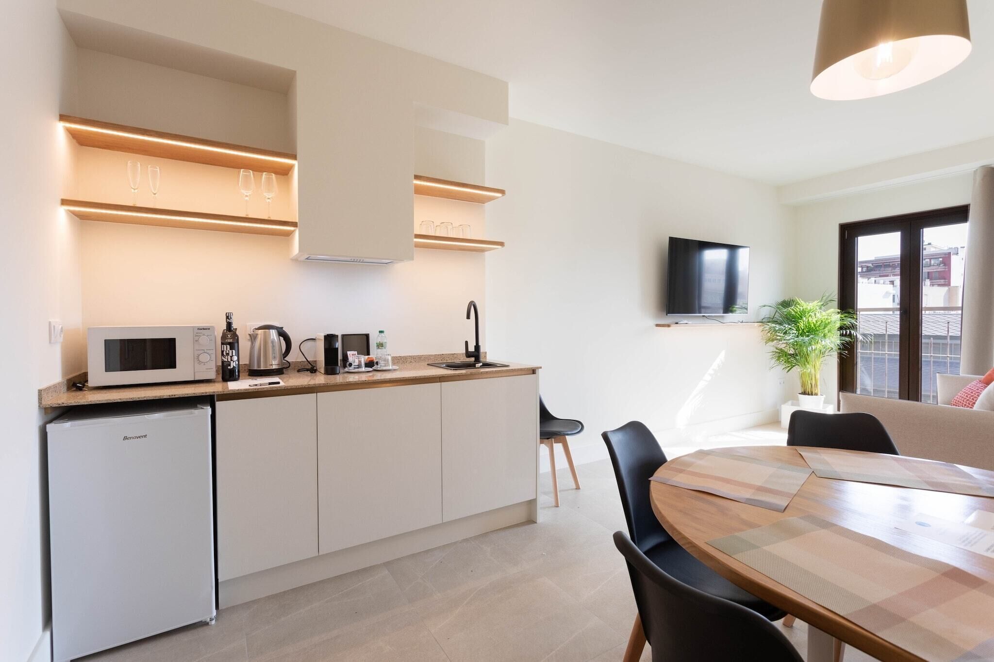 A modern kitchen with a microwave, kettle, and coffee maker, a dining area with a table and chairs, and a TV mounted on the wall.