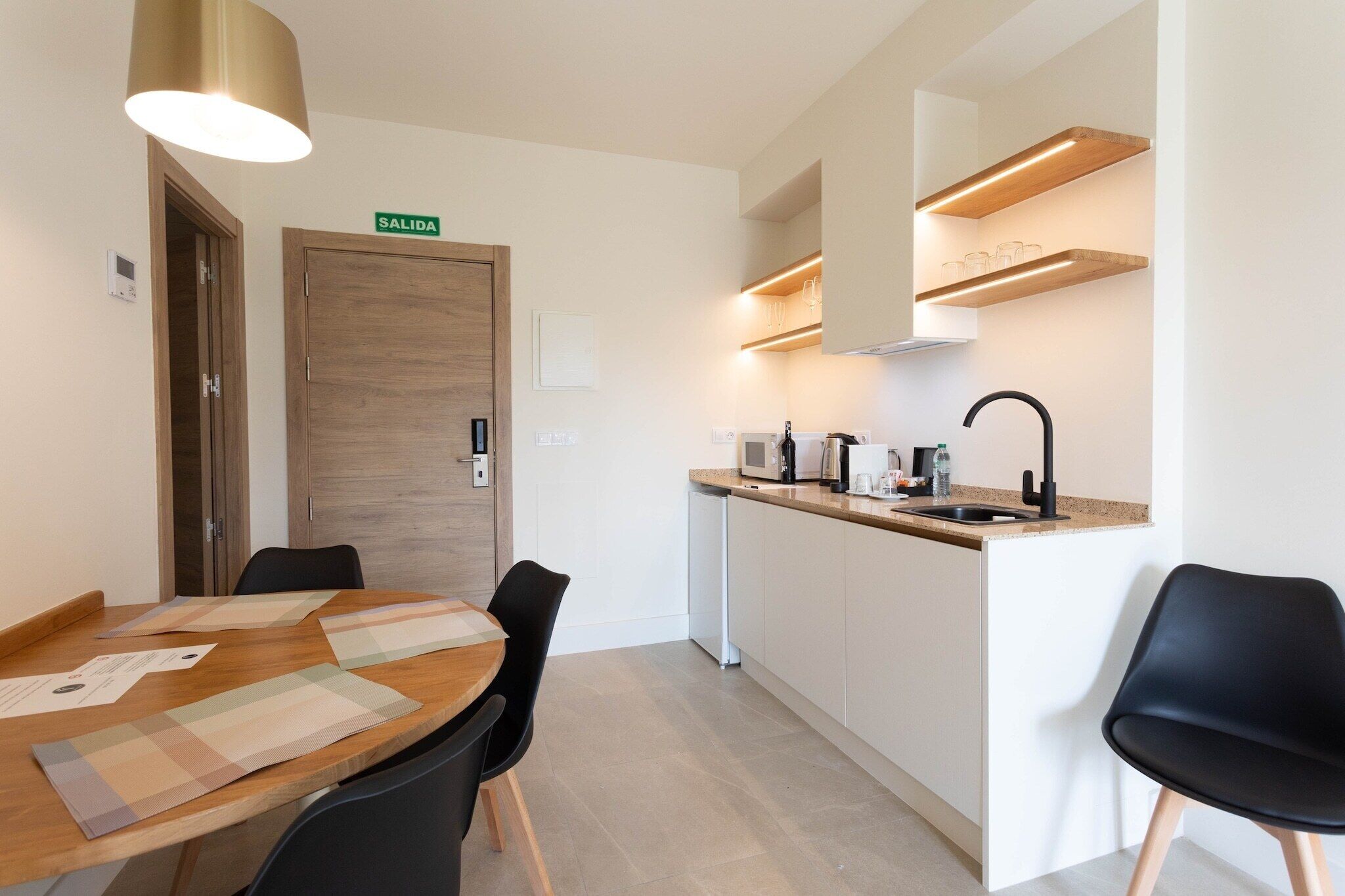 Una cucina moderna con microonde integrato, macchina del caffè e lavandino. Si vede una porta in legno con la scritta 'SALIDA' sopra, e un'area pranzo con tavolo e sedie.