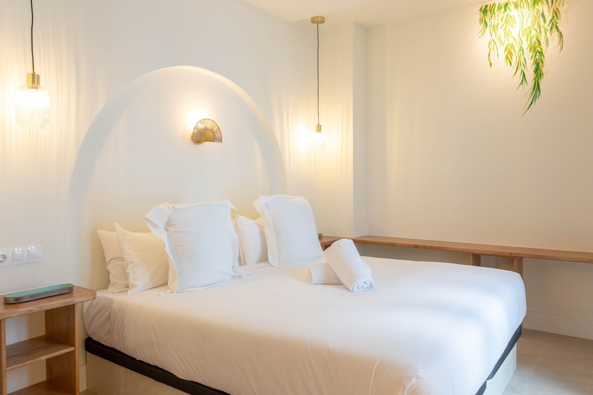 Una cama bien hecha con sábanas y almohadas blancas, una mesita de noche con una bandeja y una planta colgante.