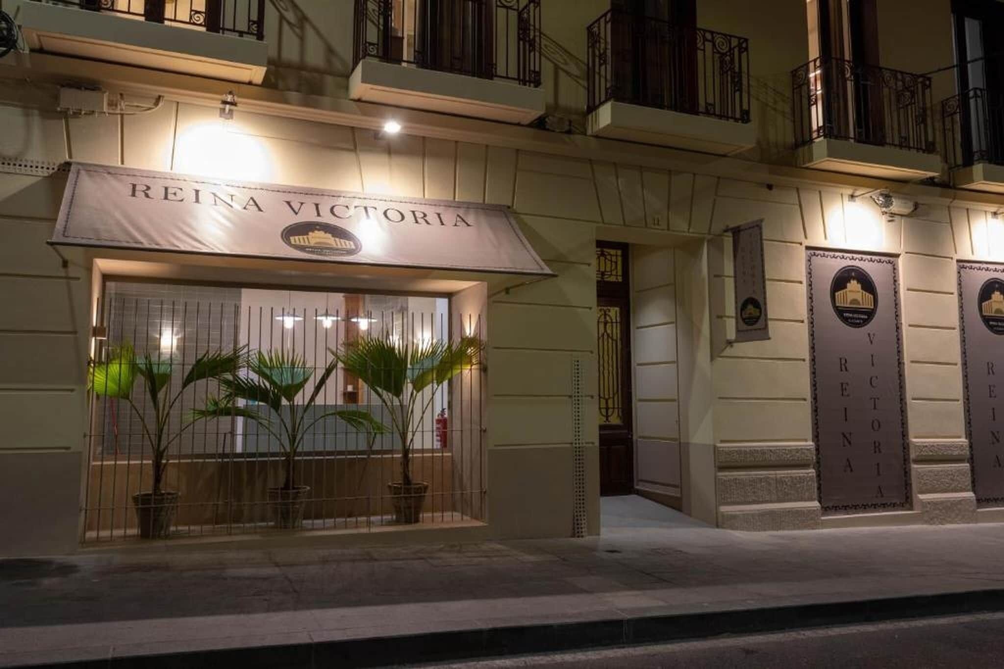 Entrada de un hotel con un letrero que dice 'REINA VICTORIA', plantas en macetas y una entrada con portones.