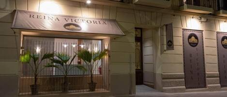 L’entrée d’un hôtel, avec une enseigne indiquant « REINA VICTORIA », des plantes en pot et une entrée fermée par un portail.