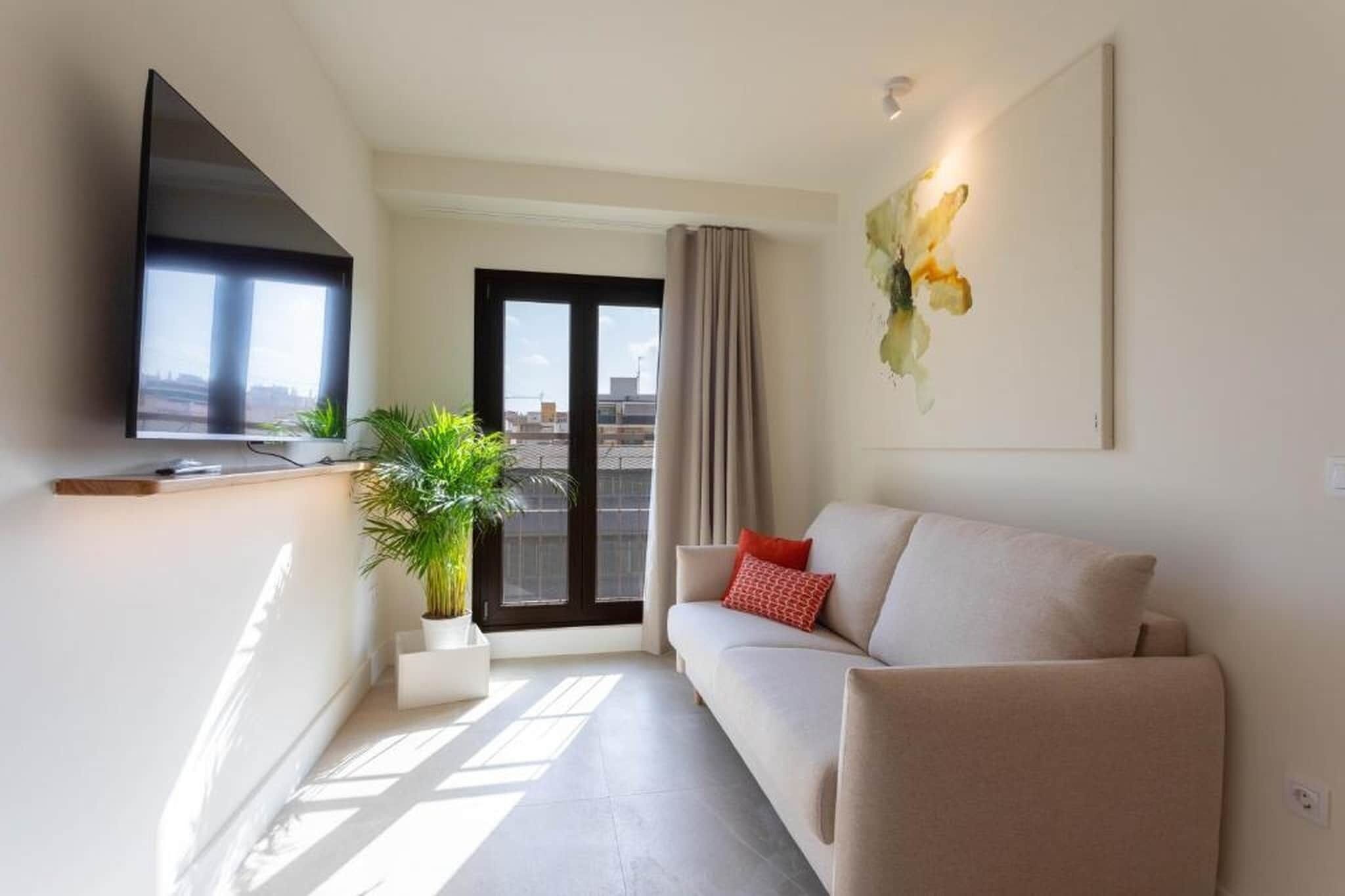 Een moderne woonkamer met een bank, een flatscreen televisie, een potplant en uitzicht op de stad door een groot raam.