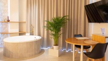 A modern bathroom with a freestanding bathtub, a potted plant, a wooden table with chairs, and a flat-screen TV mounted on the wall.