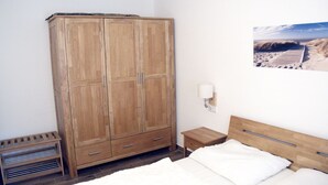 A bedroom with a wooden wardrobe, a bedside table, a nightstand, a bed with white bedding, and a wall-mounted lamp. - Vacation Rental on Nordstrand—Close to the Beach and Comfortable—Comfortable (Dranske)
