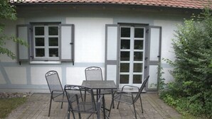 A patio with a table and chairs, a window with shutters, and a door with glass panels. - Vacation Rental on Nordstrand—Close to the Beach and Comfortable—Comfortable (Dranske)