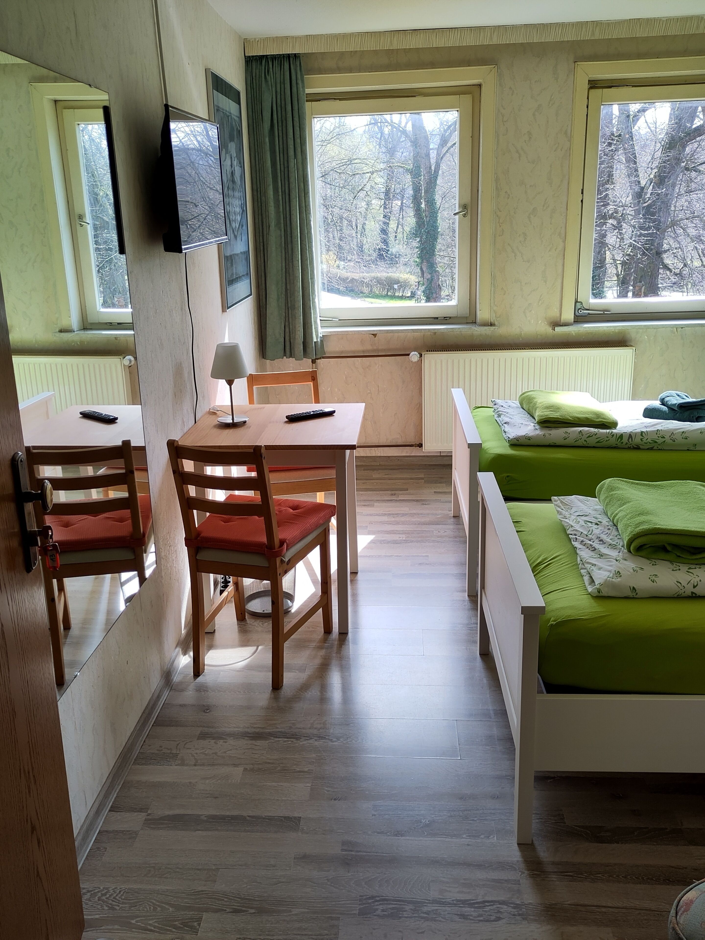 Una habitación con una mesa de madera, dos sillas, una cama con ropa de cama verde y una ventana con vistas a árboles.