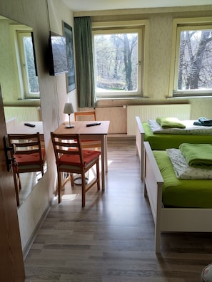 A room with a wooden table, two chairs, a bed with green bedding, and a window with a view of trees.