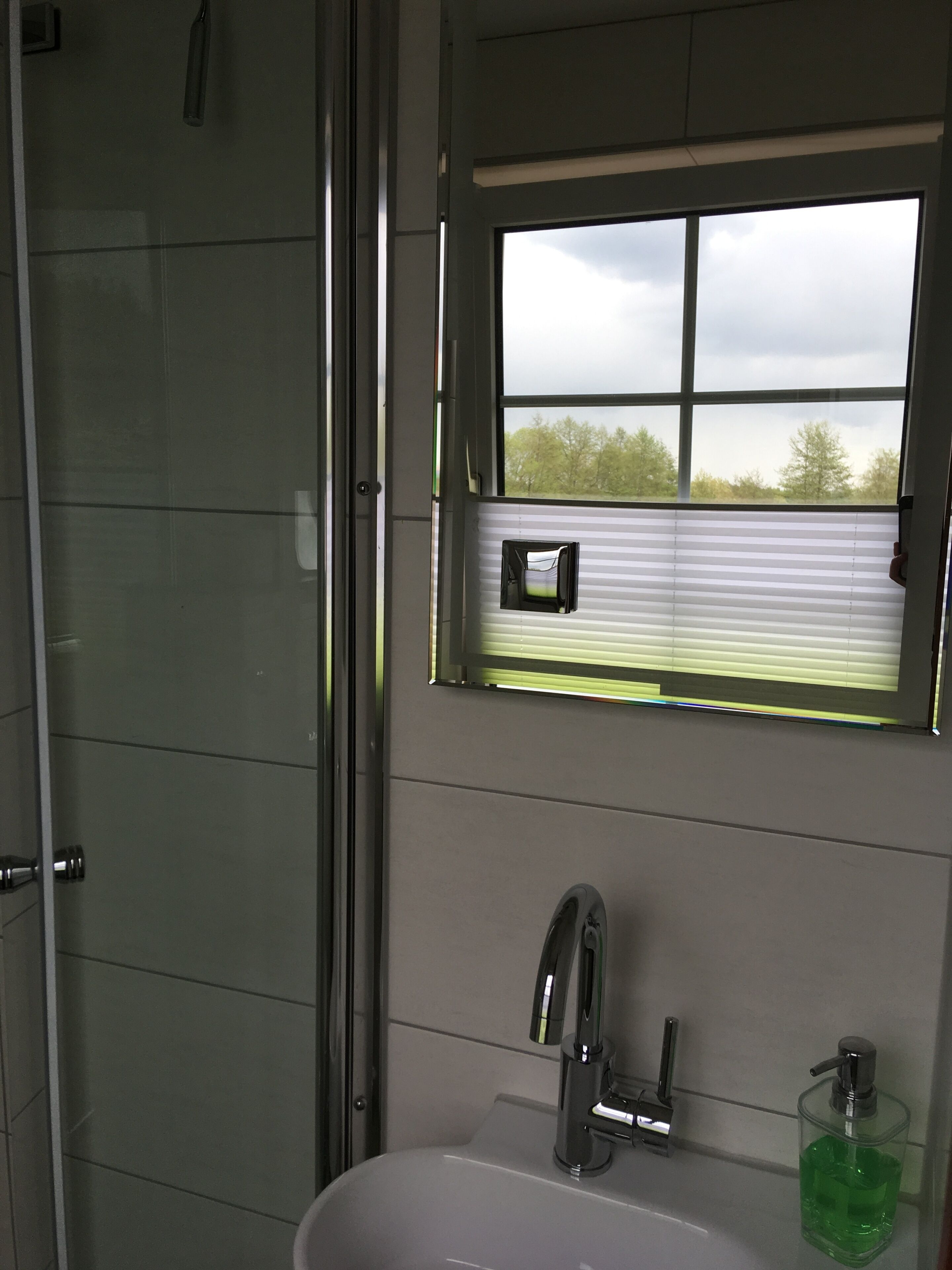 Ein Badezimmer mit Dusche, einem Fenster mit Jalousien, einem Waschbecken mit Wasserhahn und einer Seifenspender.