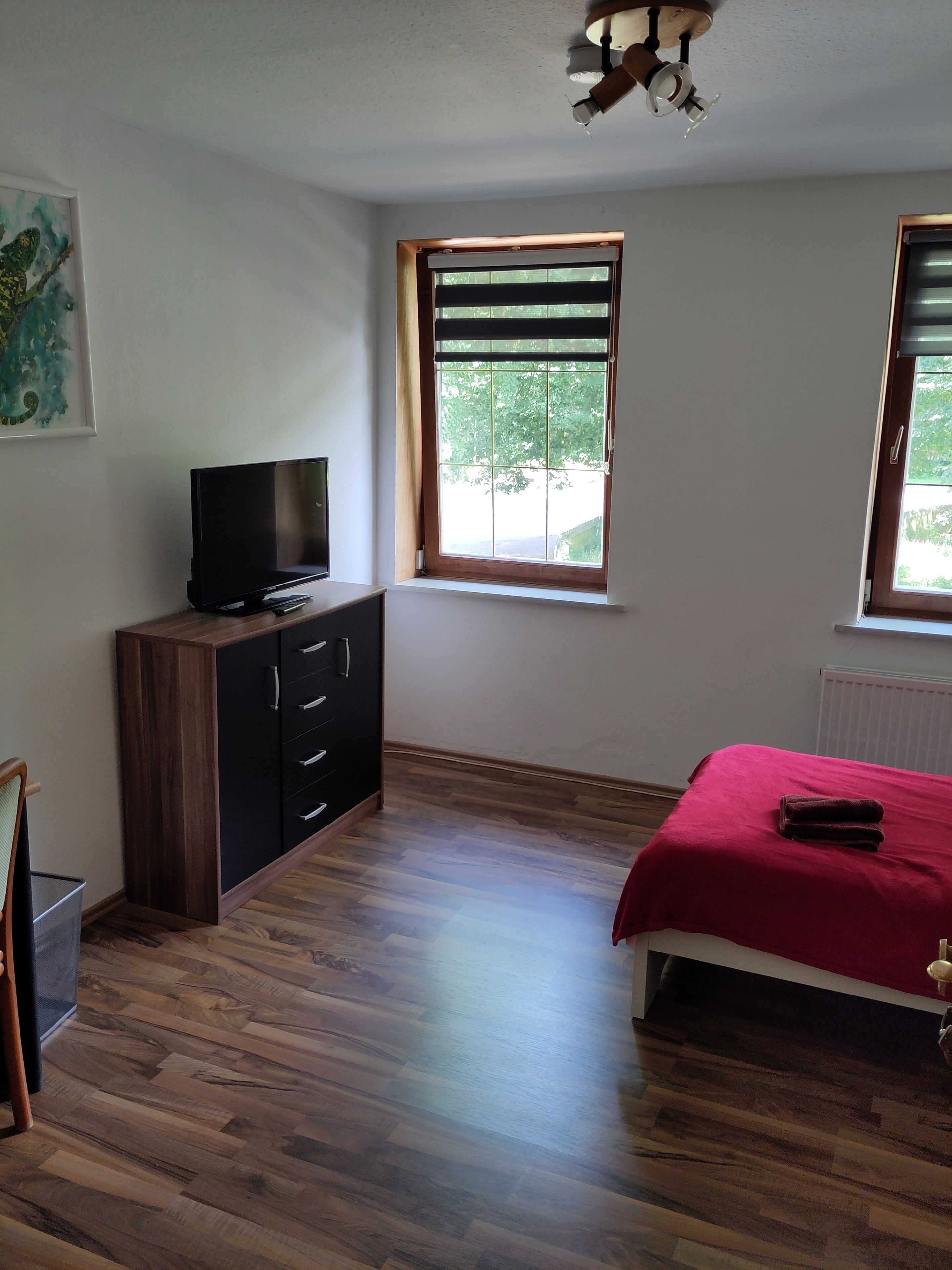Une chambre à coucher avec un lit, un téléviseur posé sur un meuble en bois, une fenêtre avec des stores et un ventilateur de plafond.