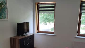 Quarto com cama, televisão sobre um armário de madeira, janela com persianas e ventilador de teto.