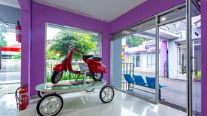 Quarto com uma scooter vermelha sobre uma mesa de vidro, um extintor de incêndio e vista para a rua com um hotel e cadeiras do lado de fora.