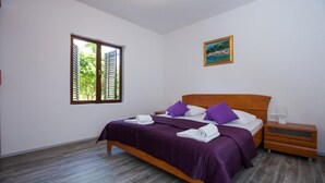 Ein Schlafzimmer mit einem hölzernen Bett, violetter Bettwäsche, einem Nachttisch mit Lampe und einem Fenster mit Rollläden.