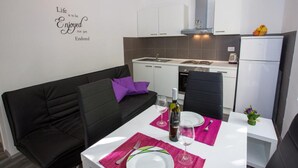 Eine kleine Küche mit weißen Schränken, ein schwarzes Sofa mit violetten Kissen und ein Esstisch für zwei Personen mit rosa Unterlagen.
