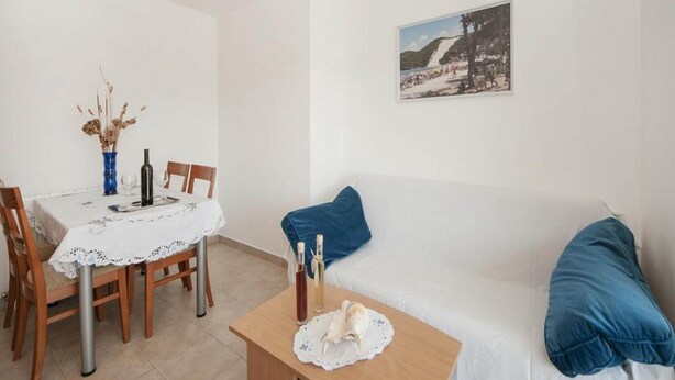 A room with a dining table, chairs, a white sofa with blue cushions, a small wooden table with a bottle and glass, and a wall-mounted picture. - Bertie's Lodge - Two Bedroom Apartment With Terrace and Sea View (Smokvica)