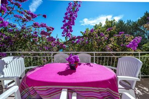 Ein runder Tisch mit rosa Tischdecke und gestreiftem Tischläufer, umgeben von weißen Stühlen, auf einem Balkon mit lila Blumen und blauem Himmel.
