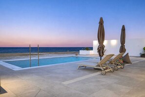 Zona junto a la piscina con sillones de playa y sombrillas, con vista a una playa y al océano.