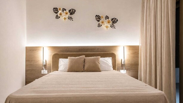 Een net opgemaakt bed met hoofdeinde, twee wandlampen en een groot raam met gordijnen.