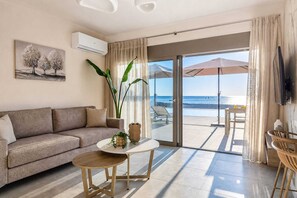 Ein modernes Wohnzimmer mit einer Couch, einem kleinen runden Tisch, einer großen Topfpflanze und Blick auf den Strand durch eine Glastür.