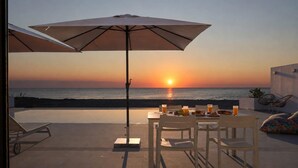 Ein Essbereich im Freien mit Blick auf den Sonnenuntergang über dem Meer, ein Tisch mit Stühlen und ein großer Sonnenschirm, der Schatten spendet.