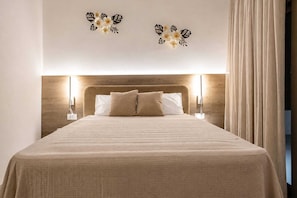 Uma cama bem arrumada com cabeceira, duas luminárias fixadas na parede e uma janela grande com cortinas.