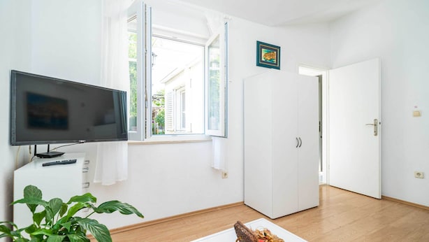 A room with a flat-screen TV, a window with curtains, a door, and a small table with a basket. - Apartment Vulicevic A2 (Dubrovnik)