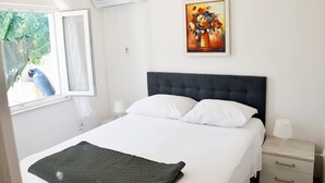 A hotel room with a bed, a nightstand, a lamp, an air conditioning unit, a window with a view of greenery, and a painting on the wall. - Apartment Vulicevic A2 (Dubrovnik)
