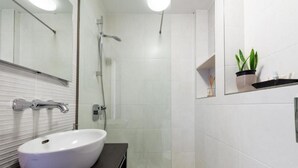 A modern bathroom with a white sink, a mirror, a shower, and a small shelf with a plant. - Apartments Meri & Karla - Two Bedroom Apartment With Balcony and Sea View (Dubrovnik)