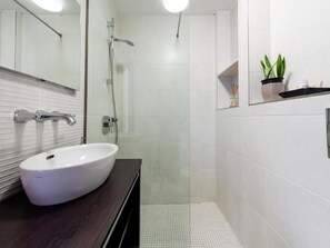 Ein modernes Badezimmer mit einem weißen Waschbecken, einem Spiegel, einer Dusche und einem kleinen Regal mit einer Pflanze.