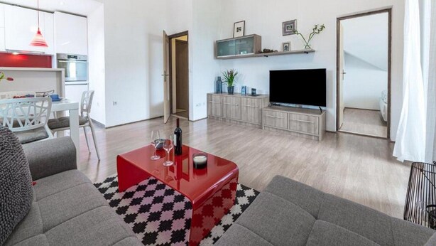 A modern living room with a red coffee table, grey sofas, a flat-screen TV, and a kitchen in the background. - Apartments Meri & Karla - Two Bedroom Apartment With Balcony and Sea View (Dubrovnik)