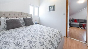 A bedroom with a bed, a nightstand, a framed picture, and a view into a living area with a sofa. - Apartments Meri & Karla - Two Bedroom Apartment With Balcony and Sea View (Dubrovnik)