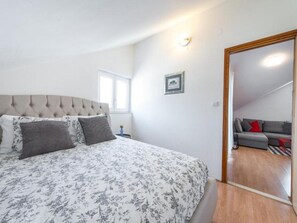 Ein Schlafzimmer mit einem Bett, einem Nachttisch, einem gerahmten Bild und Blick in einen Wohnbereich mit einer Couch.