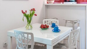 A dining area with a white table, blue chairs, a vase with red flowers, and a bowl of fruit. - Apartments Meri & Karla - Two Bedroom Apartment With Balcony and Sea View (Dubrovnik)
