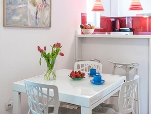 Ein Essbereich mit einem weißen Tisch, blauen Stühlen, einer Vase mit roten Blumen und einer Schale mit Obst.