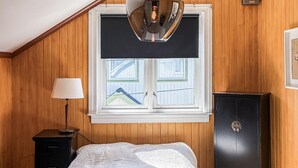 Una habitación con paneles de madera, una cama con manta blanca, un aparador, un armario negro y una ventana con cortinilla negra.