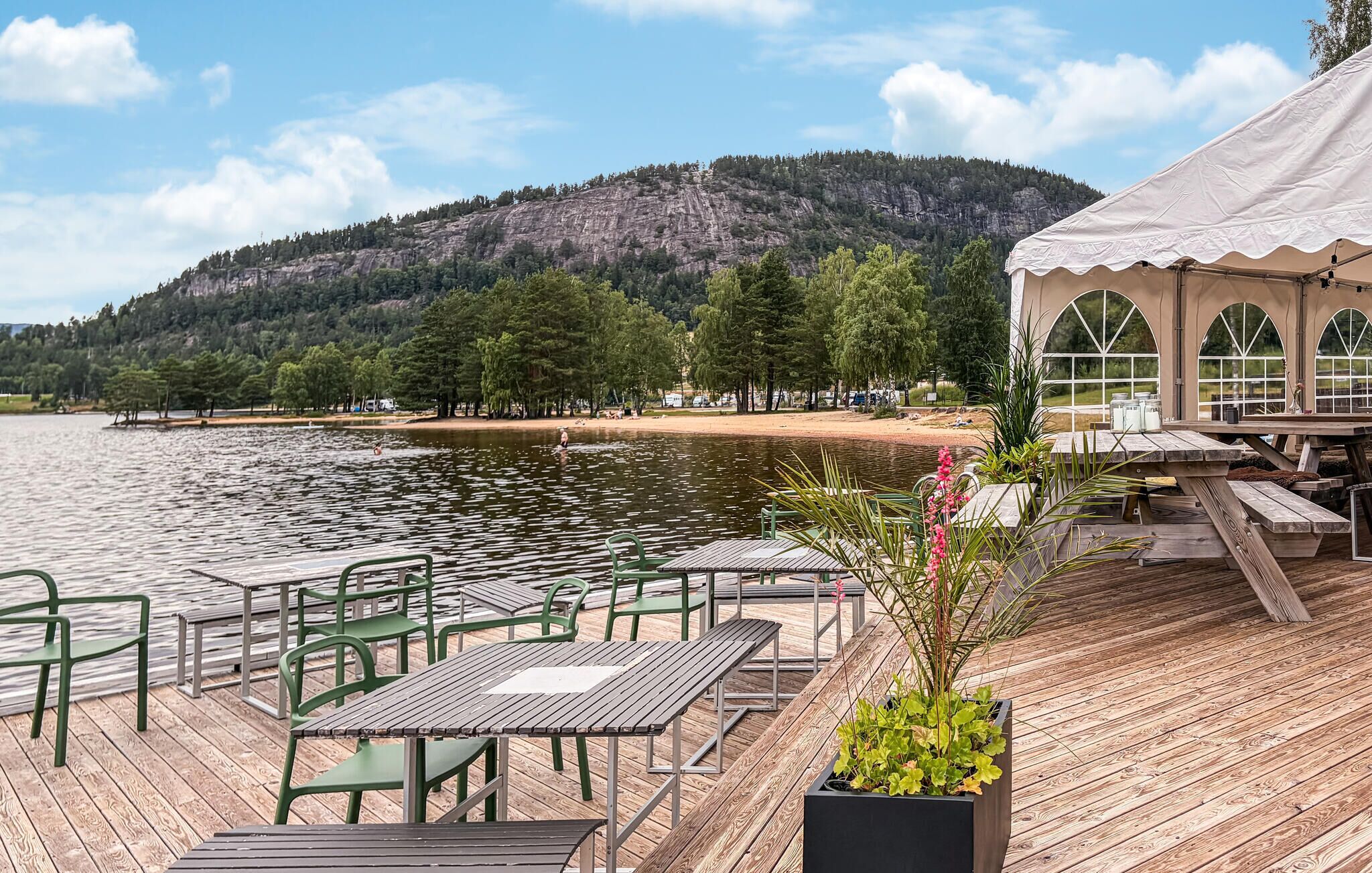 Een terras buiten aan het water met tafels en stoelen, een houten terras en uitzicht op een meer en beboste heuvels.