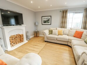 A living room with a fireplace, a sofa set, a television, and a window with curtains. - 1 The Old Smithy (Cemaes Bay)