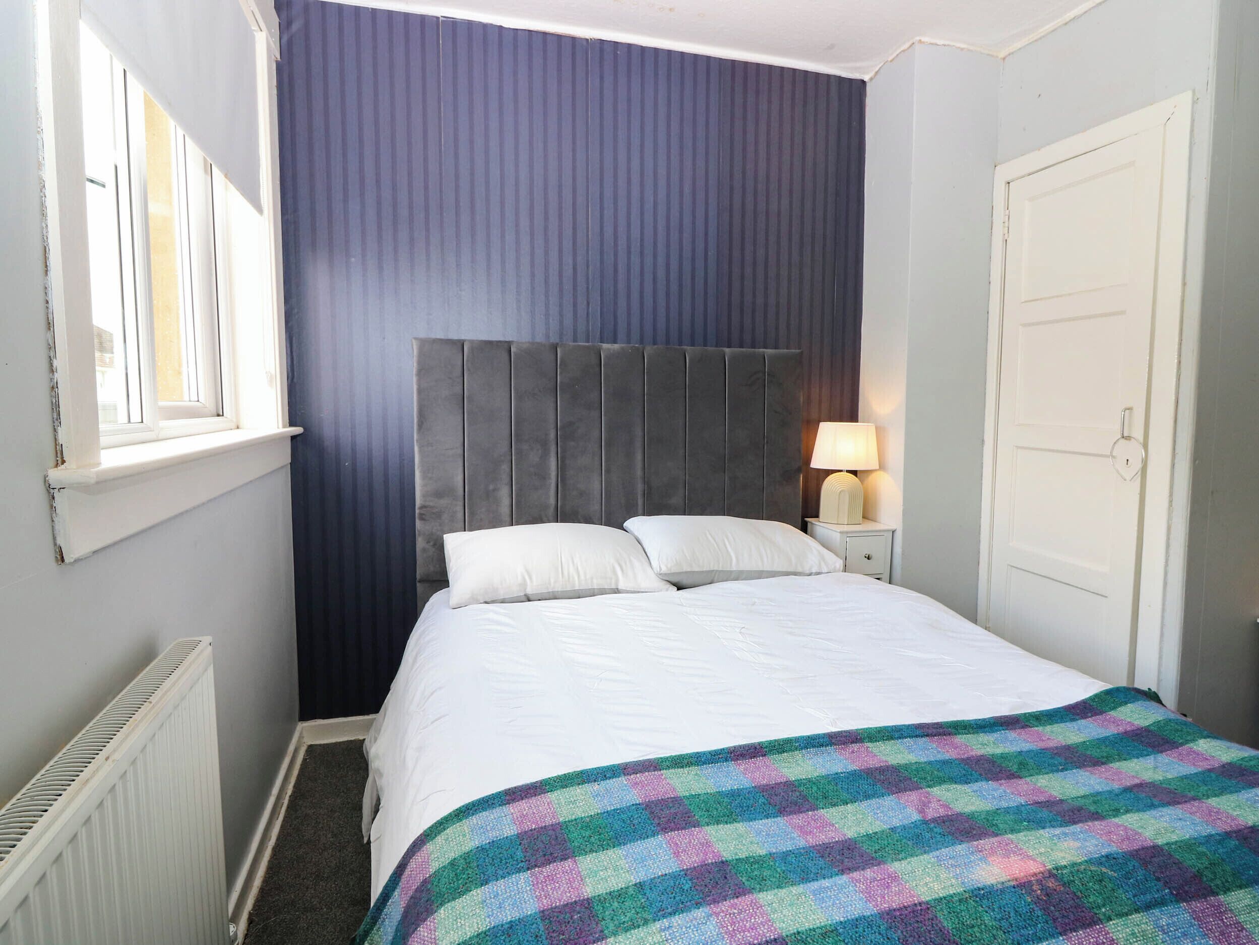 A bedroom with a bed, a nightstand, a window, a radiator, and a door.