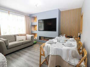 Un salón con sofá, una mesa de comedor para cuatro personas, un televisor de pantalla plana montado en la pared y una chimenea.