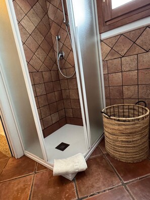 Esquina de un baño con cabina de ducha, suelo de baldosas y una cesta de mimbre.