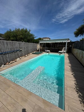 Una piscina rectangular con agua cristalina, rodeada por una terraza de madera. Se ven dos sillones de descanso y una zona de estar cubierta con una mesa.